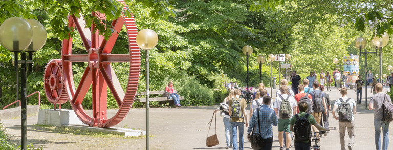 Studierende am Campus Nord Studierende laufen über den Campus. Es ist eine große rote Skulptur in Form von großen Zahnrädern zu erkennen.