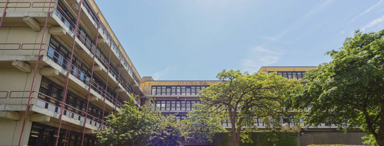 Chemiegebäude Chemiegebäude: Ein großes, breites, graues Gebäude mit Fensterfronten und Balkonen vor den Fenstern, die mit roten Metallstangen abgegrenzt werden. In der Mitte eine Treppe, auf der Menschen sitzen und links vor der Treppe Fahrräder in Fahrradständern.