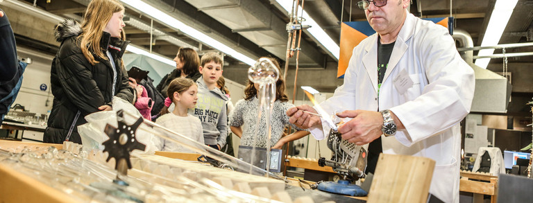 Lab A man in a white lab coat stands at a table in the lab and children stand around the table.